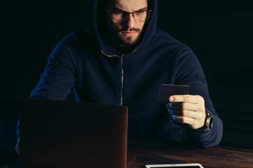 serious caucasian hacker man holding card in hand, wearing pullover, sit in the hood and studying...