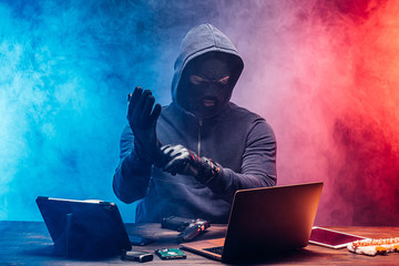 anonymous hacker male sit with laptop over smoky colorful space. professional programmer man wear black gloves not to leave fingerprints or secretly kill somebody use gun. male sit in mask in hood
