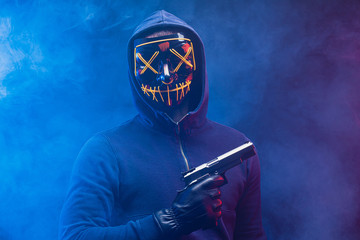 young incognito man in mask stand with gun isolated over smoky space, wearing pullover, in the hood. anonymous burglary male look at camera, engaged in cyberattack, in crime