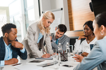 selective focus of attractive businesswoman writing in notebook near multicultural colleagues sitting at desk in conference hall
