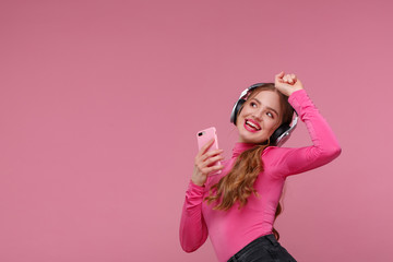 Enjoy listening to music. Beautiful young redhead woman with headphones listening music and making selfie. Funny emotional smiling girl with earphones and mobile phone on pink background.