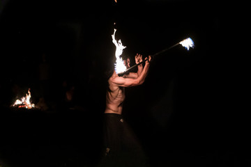 Fototapeta na wymiar A side profile view with selective focus on a muscular fire dancer performing with a flaming stick outdoors by night, details of the arm and torso muscles working