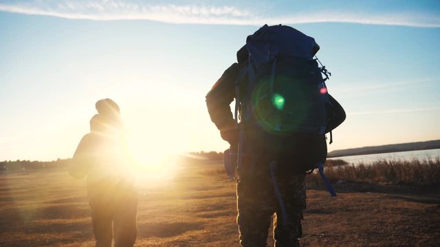 two tourists teamwork with backpacks slow motion walking go silhouette in the sunlight lifestyle glare of the sun at sunset. two men hikers climb mountains overcoming difficulties concept teamwork