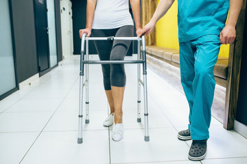 Woman using a walker, walks along the corridor of a modern rehabilitation clinic. Rehabilitologist helps patient while walking. Post-injury rehabilitation - obrazy, fototapety, plakaty