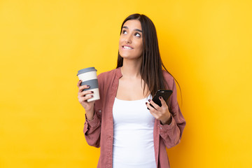 Young brunette woman over isolated yellow background holding coffee to take away and a mobile while thinking something