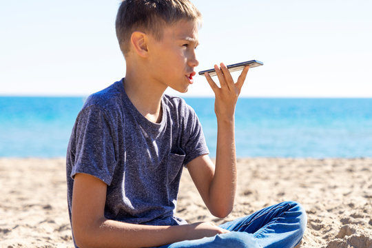 Teenager boy using a smart phone voice recognition function online, using cell phone to send a voice message on the beach