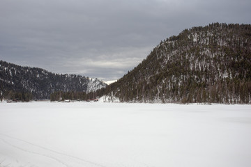 Vast frozen lake covered in fresh snow surrounded by tree covered mountains on overcast day