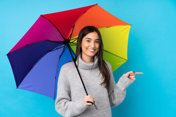 Young brunette woman holding an umbrella over isolated blue wall pointing finger to the side