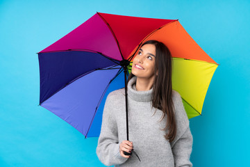 Young brunette woman holding an umbrella over isolated blue wall laughing and looking up