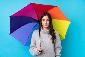 Young brunette woman holding an umbrella over isolated blue wall keeping arms crossed