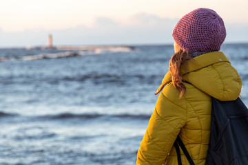 Beautiful blond caucasian girl in winter hat and bright coat looking far away at the sunset seaside