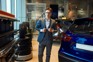 Fototapeta na wymiar Perfect car for me. young handsome man buttoning up his jacket businessman in gray checked suit standing near car at the dealership, close up photo