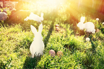 The concept of Easter holidays. Decorative Handicrafts handmade in the form of different white rabbits, located next to the painted eggs. Green grass in the background. Sunlight