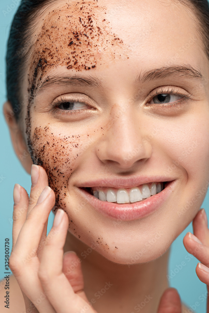 Wall mural positive woman applying coffee scrub on face, isolated on blue