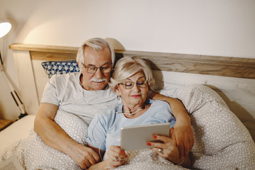 Relaxed mature couple using digital tablet while lying down in bed.