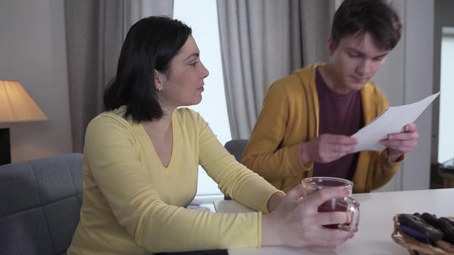 Brunette brown-eyed Caucasian woman drinking tea as teenage boy coming up and showing bad test results from college. Shocked parent getting upset of son's achievement. Disappointment, stress, trouble.