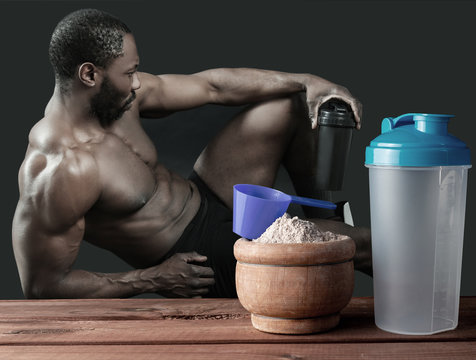 Hombre muscular con bebida proteica en el agitador sobre fondo oscuro y una mesa de madera con proteinas