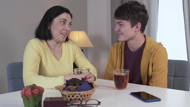 Teenage Caucasian Brunette Boy Kissing Mom On Cheek. Positive Son Spending Evening With Mother At Home. Happy Woman Talking And Adjusting Boy's Hair. Lifestyle, Family, Unity, Leisure.