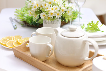 ceramic teapot and two cups on a wooden tray on the table. Plate with chopped lemon, a bouquet of daisies, plates and cutlery..