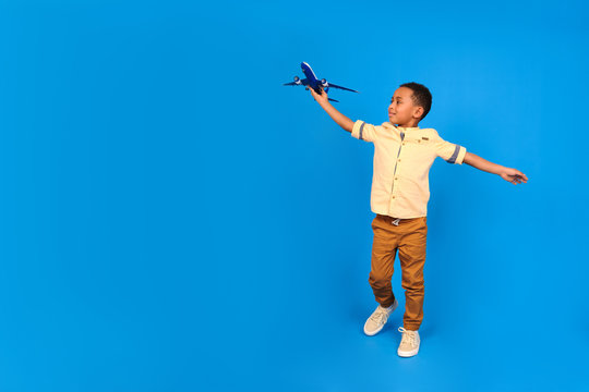 Happy African-American boy plays with a toy plane, puts his hands aside, imagines himself flying in a plane against a blue background. Family travel and tourism, summer holidays concept.