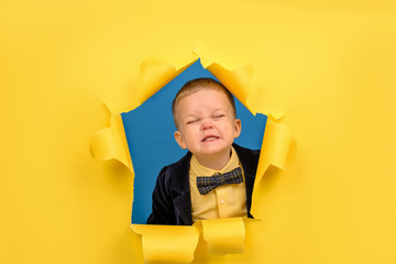 Boy looks out of hole of torn yellow paper with torn edges in jacket and bow tie and shows good positive emotion on his face. Concept of an advertising poster and good holiday mood with place for text