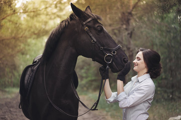 Beautiful girl jockey rider with a black horse, dressed in a light shirt in a green forest Park....