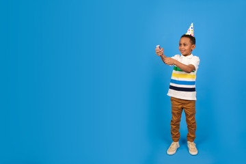 Fototapeta na wymiar African American boy blows up flapper holding her in his hand dressed in white polo and party cap stands on blue studio background. Birthday concept and holidays party concept with place for text.