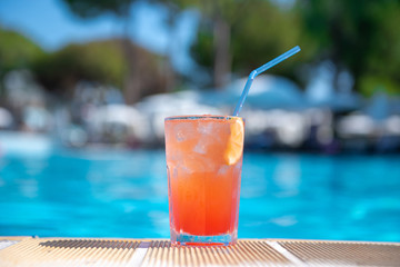 Fresh alcoholic red cocktail sex on beach with ice in glass on edge of pool in summer weather against background washed up pool and recreation area. Concept good holiday and travel, place for text.