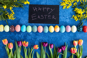 Colorful Easter eggs and tulips on dark blue background. On blackboard is written happy easter. Holiday decorations, Easter concept background.