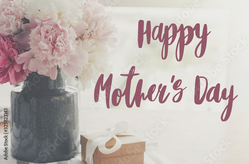 Happy mother's day lettering on pink and white peonies in vase and gift box with on rustic table in light, space for text. Happy mother's day text. Floral greeting card or poster