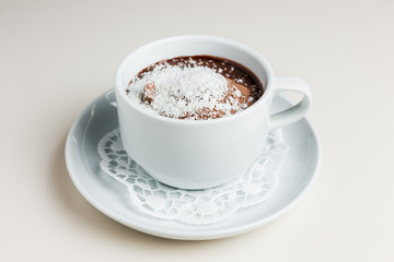 A small white cup of dark hot chocolate with chocolate ice-cream and coconut flakes on a white saucer and a napkin