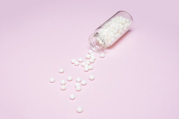 Homeopathic globules scattered from a clear glass bottle on light pink background, alternative homeopathy medicine concept