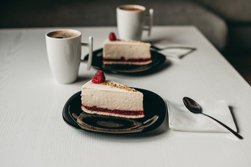 two pieces of raspberry cheesecake dessert on a black plate with coffee in a white cup on a white wooden table in a cafe