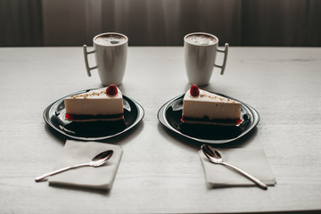 two pieces of raspberry cheesecake dessert on a black plate with coffee in a white cup on a white wooden table in a cafe