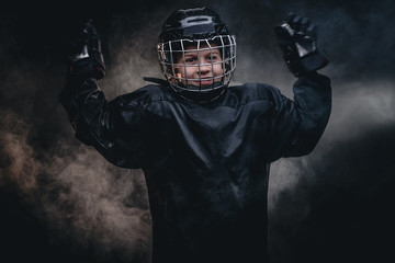 Young blonde sporty boy, ice hockey player, posing in a dark studio for a photoshoot, wearing an ice-skating uniform while holding his arms up and showing goalkeeper gloves