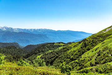 The view from the height of a green mountain valley surrounded by high mountains. Snow-capped mountain peaks on the horizon.