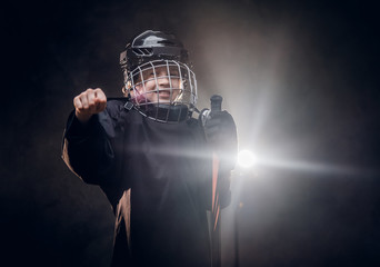 Obraz na płótnie Canvas Young blonde sporty boy, ice hockey player, posing in a dark studio for a photoshoot, wearing an ice-skating uniform, helmet, hockey stick, showing a gesture with his hand and smiling