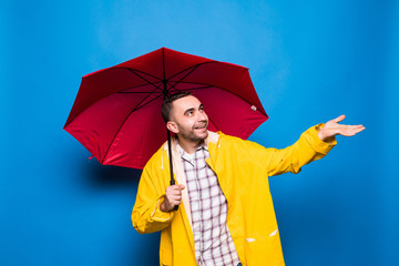 Young handsome bearded man in yellow raincoat with red umbrella isolated over blue background