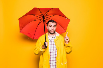 Young handsome bearded man in yellow raincoat with red umbrella have idea isolated over orange background