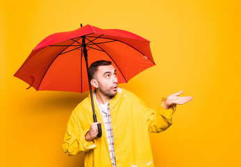 Young handsome bearded man in yellow raincoat with red umbrella trying to see if its raining isolated over orange background