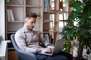 Young businessman sitting in armchair indoors in office, using laptop.