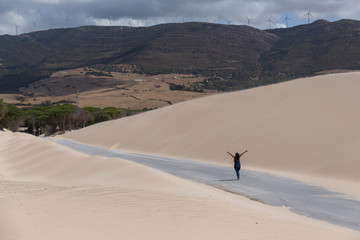 woman with arms raised walks along the road between sand dunes, in the background you can see the mountains
