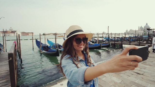 Beautiful girl in hat, sunglasses poses with smile for selfie against gondolas