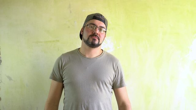 A man in a cap with a beard is standing against the wall. Serious look. Ganster in the area. The representative of a criminal gang. Military T-shirt.