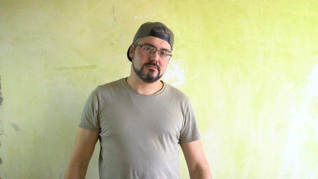 A man in a cap with a beard is standing against the wall. He directs his hands at the viewer and calls to him. Serious look. Ganster in the area. The representative of a criminal gang. Military T-shir