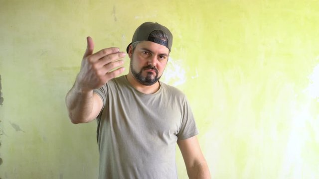A man in a cap with a beard is standing against the wall. He directs his hands at the viewer and calls to him. Serious look. Ganster in the area. The representative of a criminal gang. Military T-shir