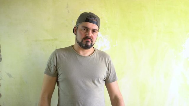 A man in a cap with a beard is standing against the wall. He directs his hands at the viewer and calls to him. Serious look. Ganster in the area. The representative of a criminal gang. Military T-shir