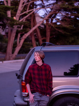 Teenager leaning against SUV looking off frame at sunset