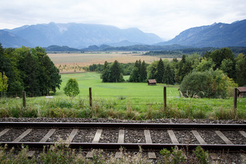 Railroad tracks and pasture fence in the foreground with a view of the moor landscape with haystacks and fir trees as well as the mountains of the Alps in the background