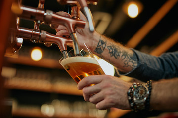 Fototapeta na wymiar Tattooed caucasian barman pouring beer while standing in pub. Selective focus on hand.